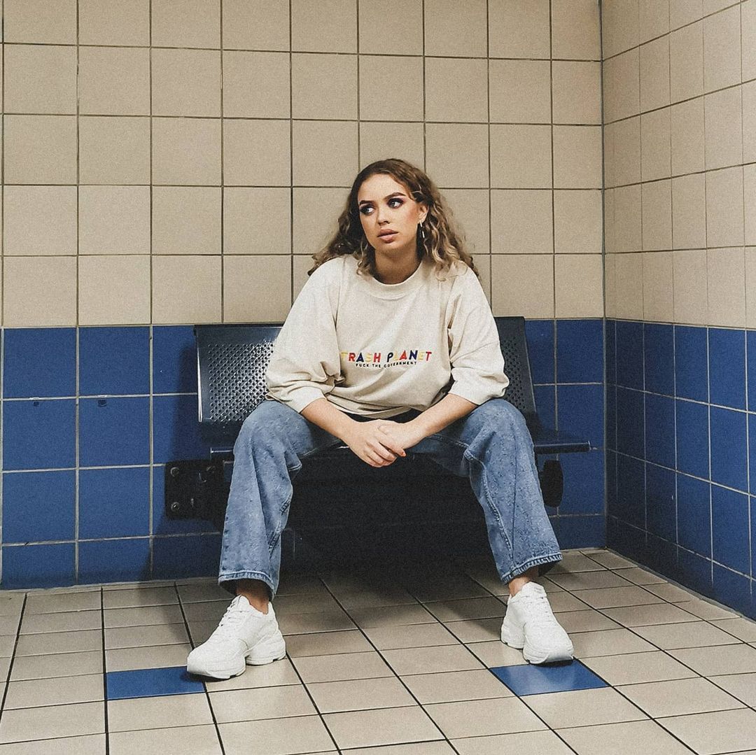 A streetwear style woman wearing an oversized trash plant t-shirt sat on a bench wearing chunky white vegan sneakers