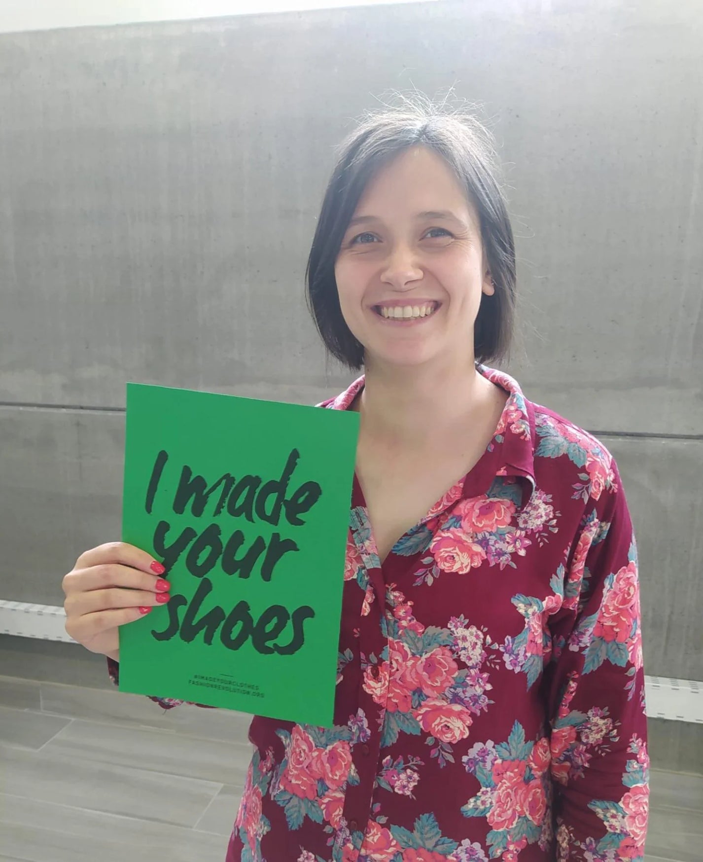 A dark haired woman from Trash Planet's footwear manufacturing facility holds up a green Fashion Revolution I Made Your Shoes Poster