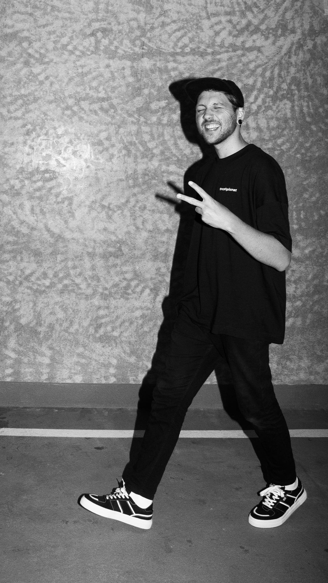 Man in an all-black streetwear fit wearing an oversized trashplanet t-shirt, black denim jeans and black cap in a pair of trash planet's recycled vegan skater style sneakers