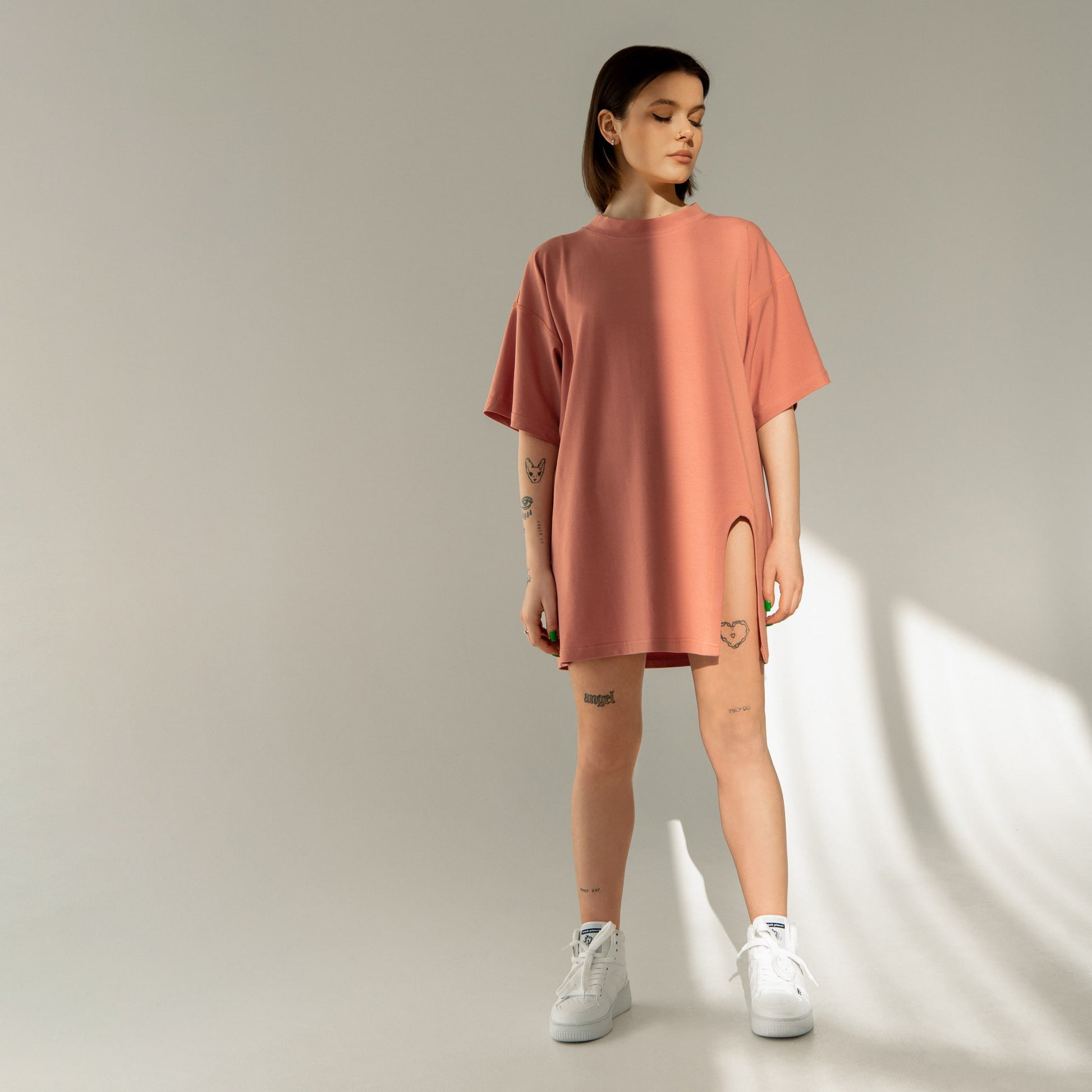 A cool streetwear style dark-haired tattooed woman is wearing an oversized coral t-shirt with a leg slit in a studio, and also wears Trash Planet's all-white vegan high-top sneakers, which are made from food waste.