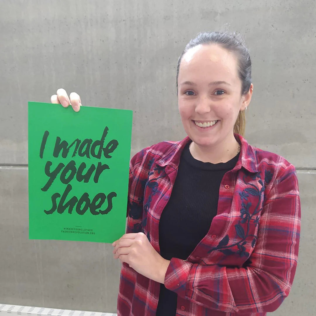 A fair haired woman from Trash Planet's footwear manufacturing facility holds up a green Fashion Revolution I Made Your Shoes Poster