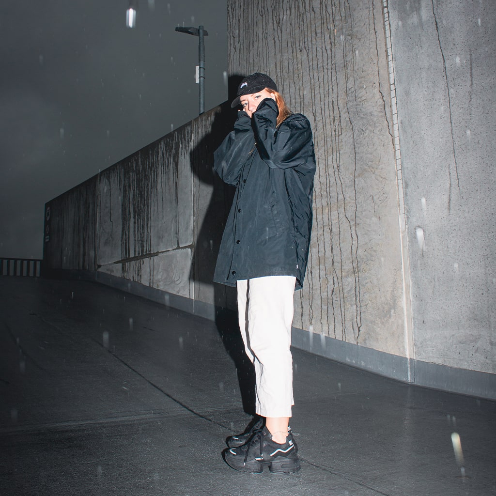 A streetwear style woman covers her face, wearing a black coach jacket, stussy cap, white jeans, and chunky split-sole black recycled vegan sneakers stood at the top of a rooftop carpark.