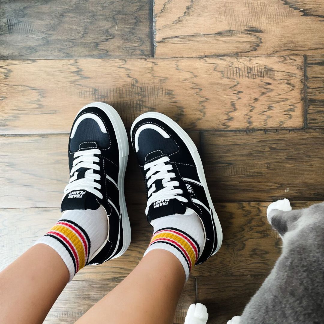 View from above of someones legs stood on a wooden floor next to a grey and white cat - the person is wearing black vegan skater style recycled sneakers with striped tube socks in autumnal colours