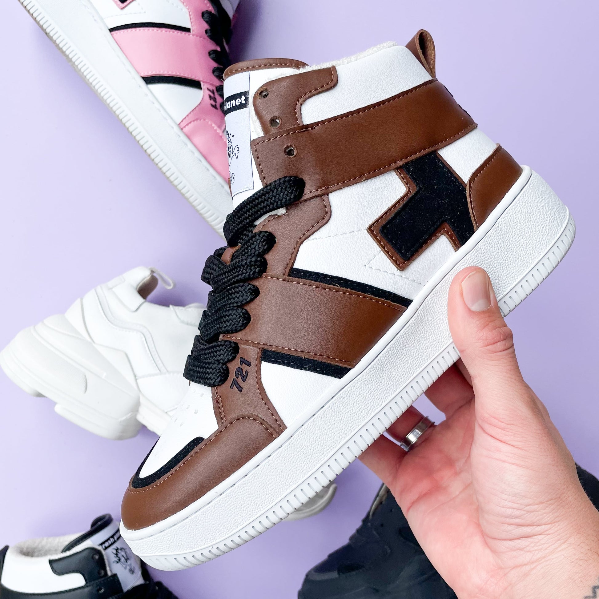A hand holds out a dark brown vegan high-top sneaker with streetwear style black and white details, which is made with recycled trash against a pastel lilac background.