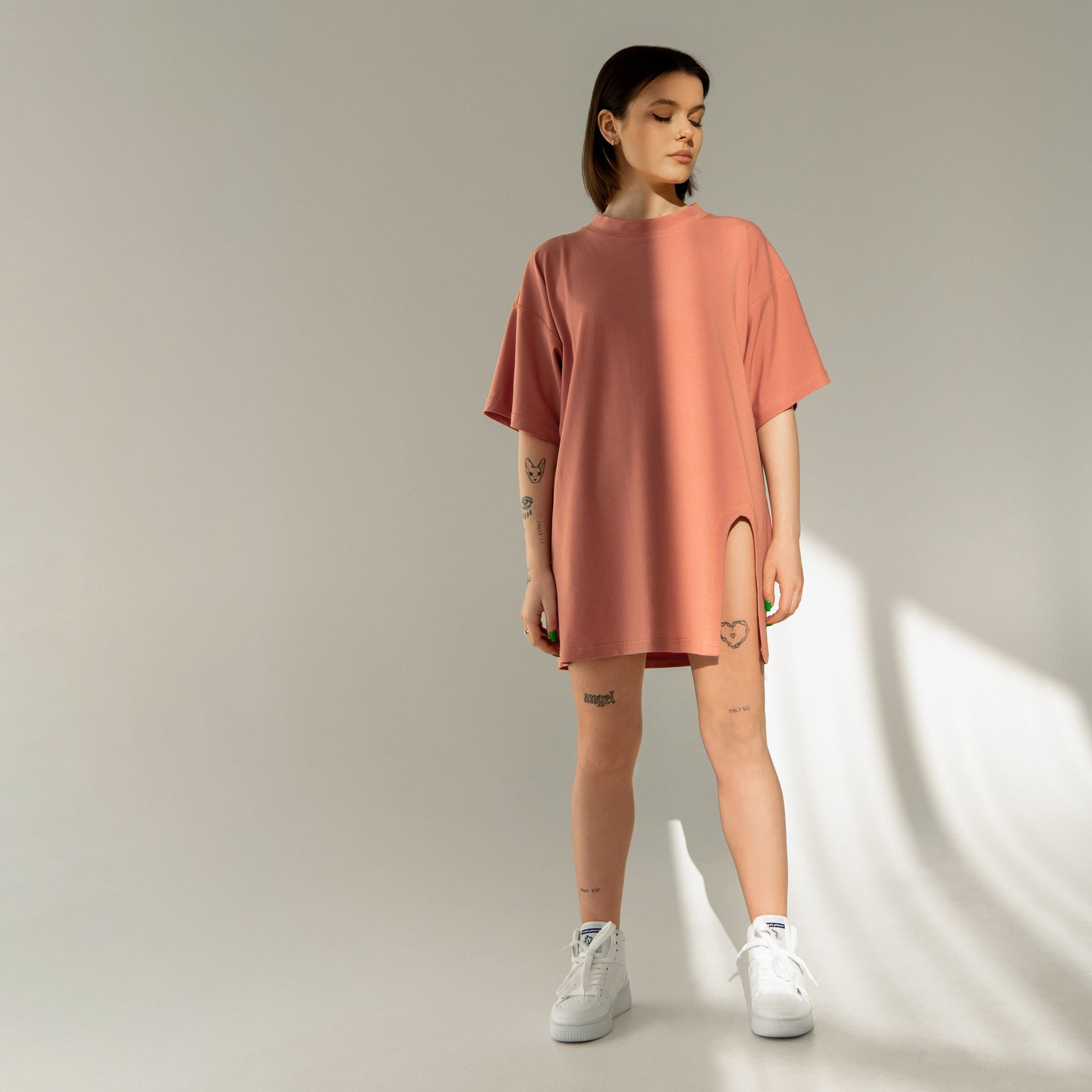 A cool streetwear style dark-haired tattooed woman is wearing an oversized coral t-shirt with a leg slit in a studio, and also wears Trash Planet's all-white vegan high-top sneakers, which are made from food waste. 