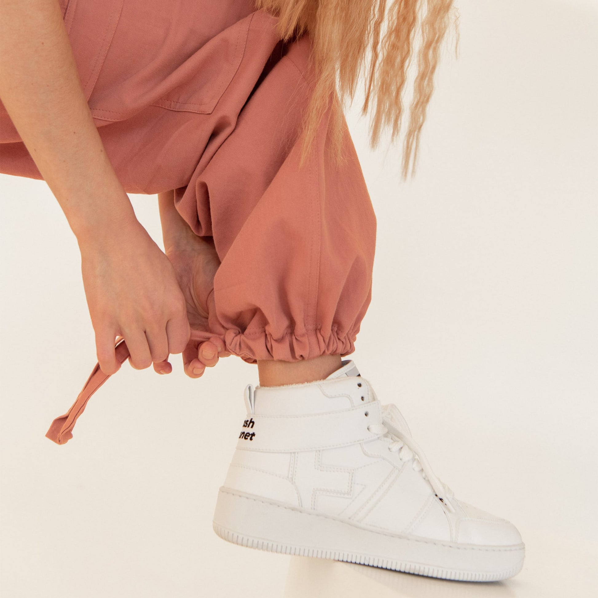 A woman kneels down to tie up a lace at the back of her streetwear style coral tracksuit to tighten it and is wearing Trash Planet's Aster 721 all-white vegan high-top sneakers.