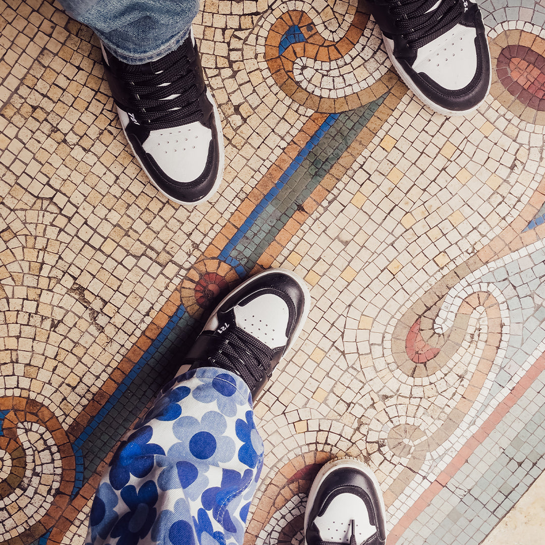 A birds eye view of two people's feet on a Roman style patterned mosaic floor; the people are wearing Trash Planet's 'Aster 721' vegan high-top sneakers made from recycled materials.