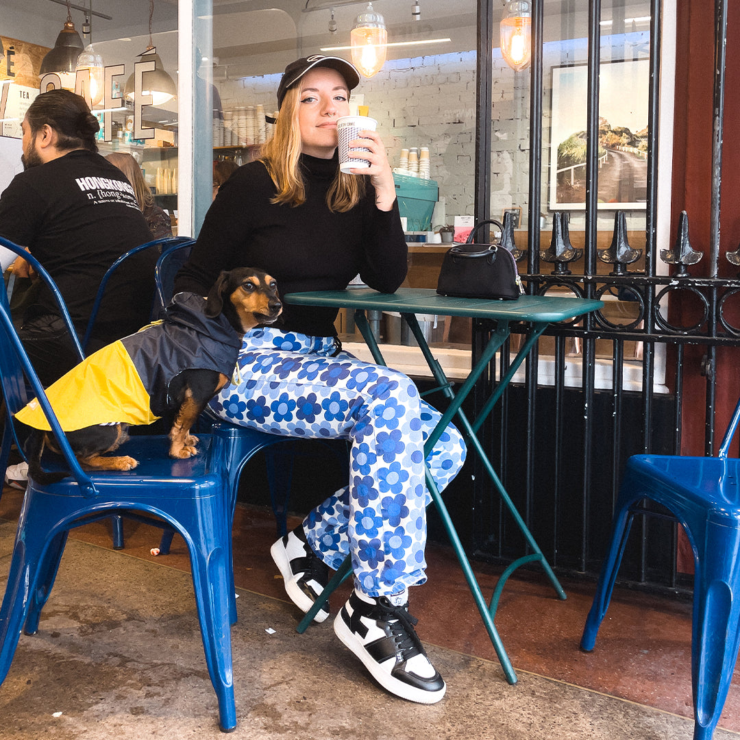    A cool streetwear style woman sat outside Society cafe on a blue metal chair drinks a coffee next to a sausage dog, which is wearing a yellow and blue coat. The woman is wearing a black and white streetwear style cap, blue and white floral patterned jeans and black and white cool streetwear vegan high-top sneakers made from waste fruit and plants.