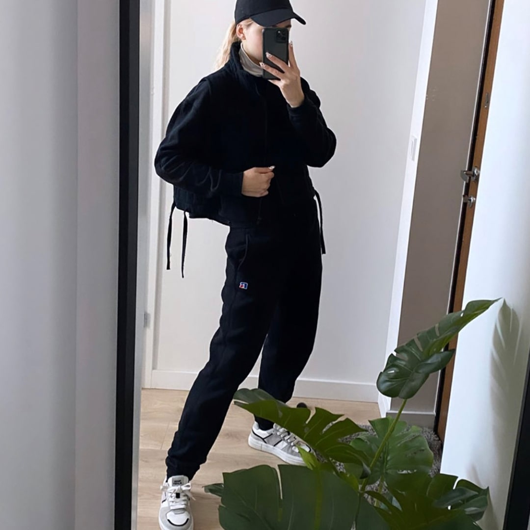A woman stood in her house in front of a mirror takes a selfie wearing a streetwear style fleece, tracksuit and black cap. She is also wearing the grey, black, and white Franco sneakers which are made from recycled trash and ocean plastic.