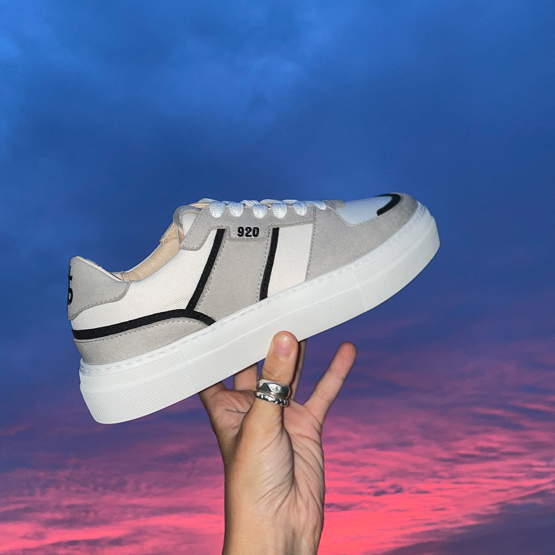 A hand with two recycled silver rings on the thumb holds up a grey, white, and black coloured vegan sneaker against a deep blue and purple sunset