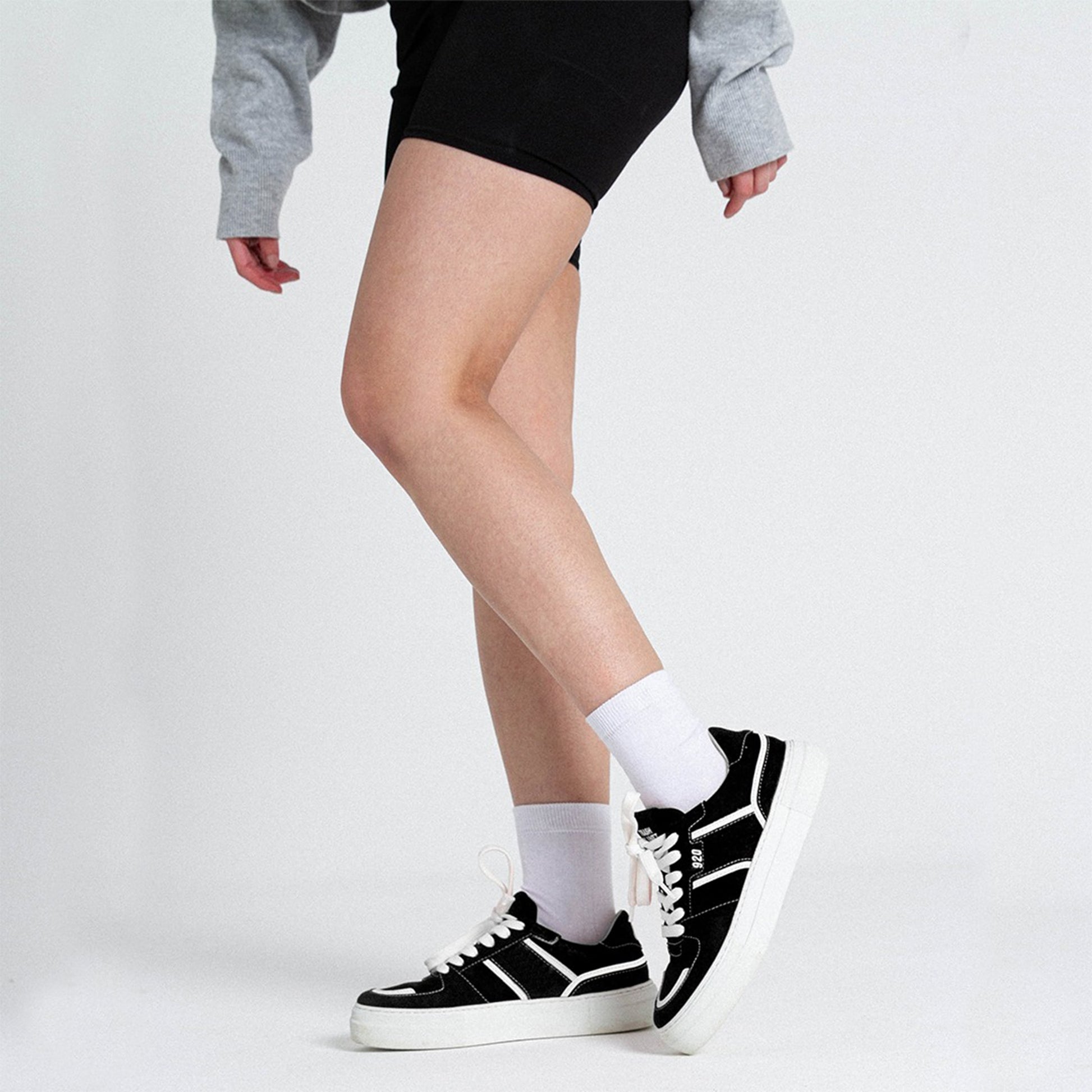 A blonde woman wearing black and white recycled vegan sneakers made from recycled trash with a loose cut grey sweater on and sporty black shorts.