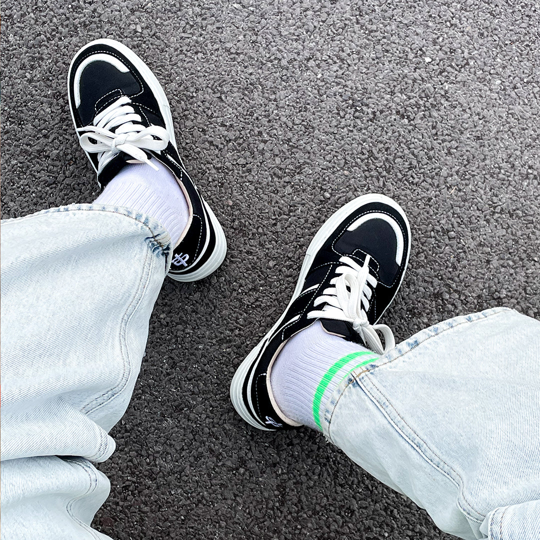 Legs on a concrete floor in a pair of black skater-style streetwear vegan sneakers with stripey tube socks in neon green and baggy blue denim jeans