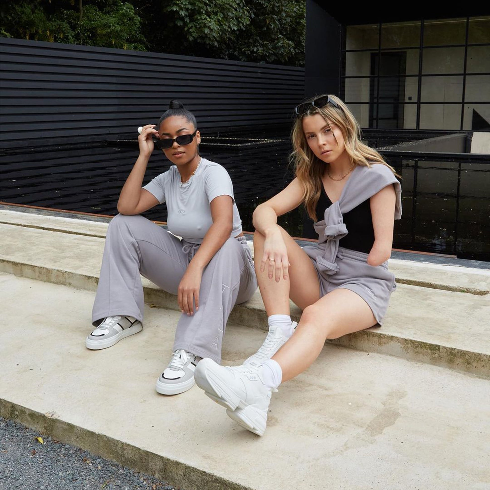 Two women sat outside of the Barbican wearing athleisure style streetwear clothes in all-grey and black sunglasses wear recycled vegan chunky platform sneakers called Billie by Trash Planet