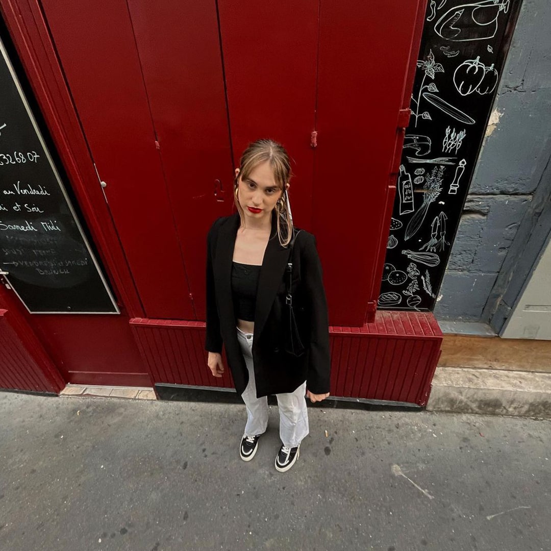 A woman stood against a red restaurant in France wearing a black blazer with a black top, white jeans, and black and white streetwear style skater vegan sneakers