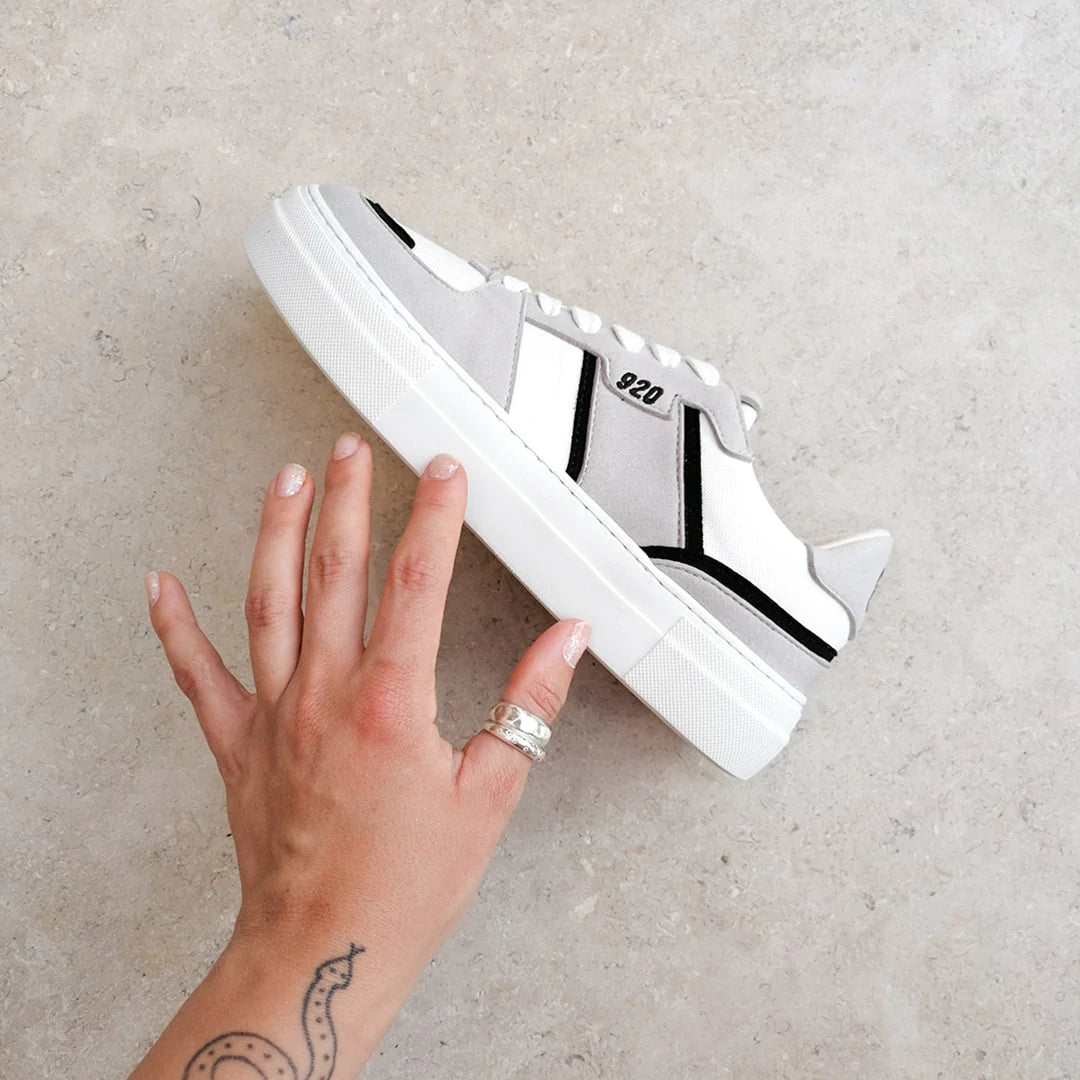 A tattooed hand with a silver ring on it holding a recycled vegan sneaker in grey, white and black against a clean granite stone backdrop